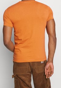 Krátký rukáv oranžové tričko vyrobené z bavlny, s kulatým výstřihem a přiléhavým střihem. Nosené s hnědými manšestrovými kalhotami.