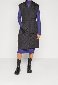 Vadderad svart väst med breda ärmar, ett knytbälte och sidofickor, över en lila klänning. Modell som bär svarta ankelboots.