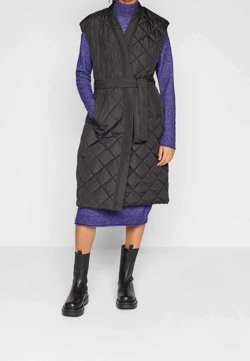 Vadderad svart väst med breda ärmar, ett knytbälte och sidofickor, över en lila klänning. Modell som bär svarta ankelboots.