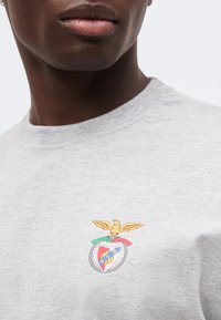 Κοντινό πλάνο ενός ατόμου που φοράει ανοιχτό γκρι πουκάμισο με πολύχρωμο έμβλημα αετού και ασπίδα στο στήθος.