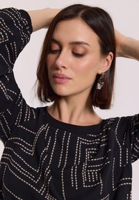 Czarny top z beżowym wzorem geometrycznym; posiada długie rękawy i okrągły dekolt. Modelka ma fryzurę bob i nosi metaliczne kolczyki.