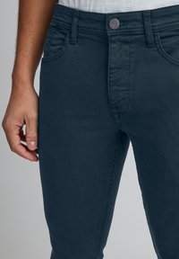 Temno modre kavbojke s tesnim krojem, z zapiranjem na gumbe, sprednjimi žepi in mehko teksturo. Roka na boku.