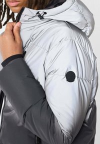 Gewatteerd jack met een grijs-zilveren kleurenschema; voorzien van een capuchon, ritssluiting en een cirkelvormig logo-accent op de schouder.