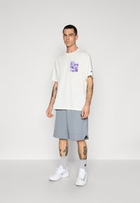 White t-shirt with purple graphic pocket, paired with light gray shorts. Model wearing white sneakers and gray socks, standing against a plain background.