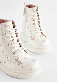 Bottes blanches de cheville avec un motif floral, dotées de lacets, d'une fermeture éclair latérale et d'une semelle en caoutchouc texturée. Doublure rose pâle ajoutant un contraste.