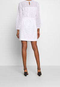 Vestido blanco con mangas largas, tejido de ojal, cintura ceñida con cinturón y detalle de abertura en la parte posterior. Termina por encima de la rodilla, con una textura suave.