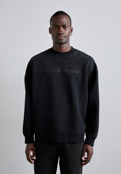 Sweatshirt noir avec des détails usés et "WON HUNDRED" en relief, doté d'un col rond et de manches longues, associé à un jean foncé.