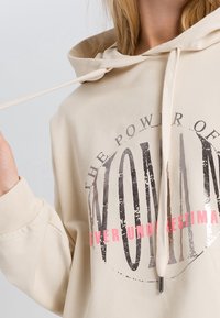Light beige hoodie with a front graphic reading “THE POWER OF WOMAN” in black and pink text. Features a drawstring and soft, textured fabric.