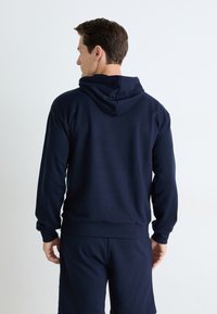 Haut en coton bleu marine avec capuche à cordon, poignets et ourlet côtelés. Présente une texture lisse et une coupe décontractée. Porté par-dessus un short assorti.