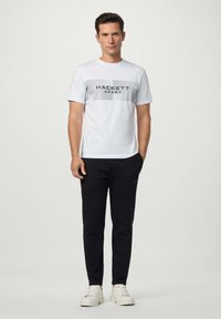 Hombre de pie vistiendo una camiseta deportiva blanca de Hackett, pantalones negros y zapatillas blancas, contra un fondo claro y liso.