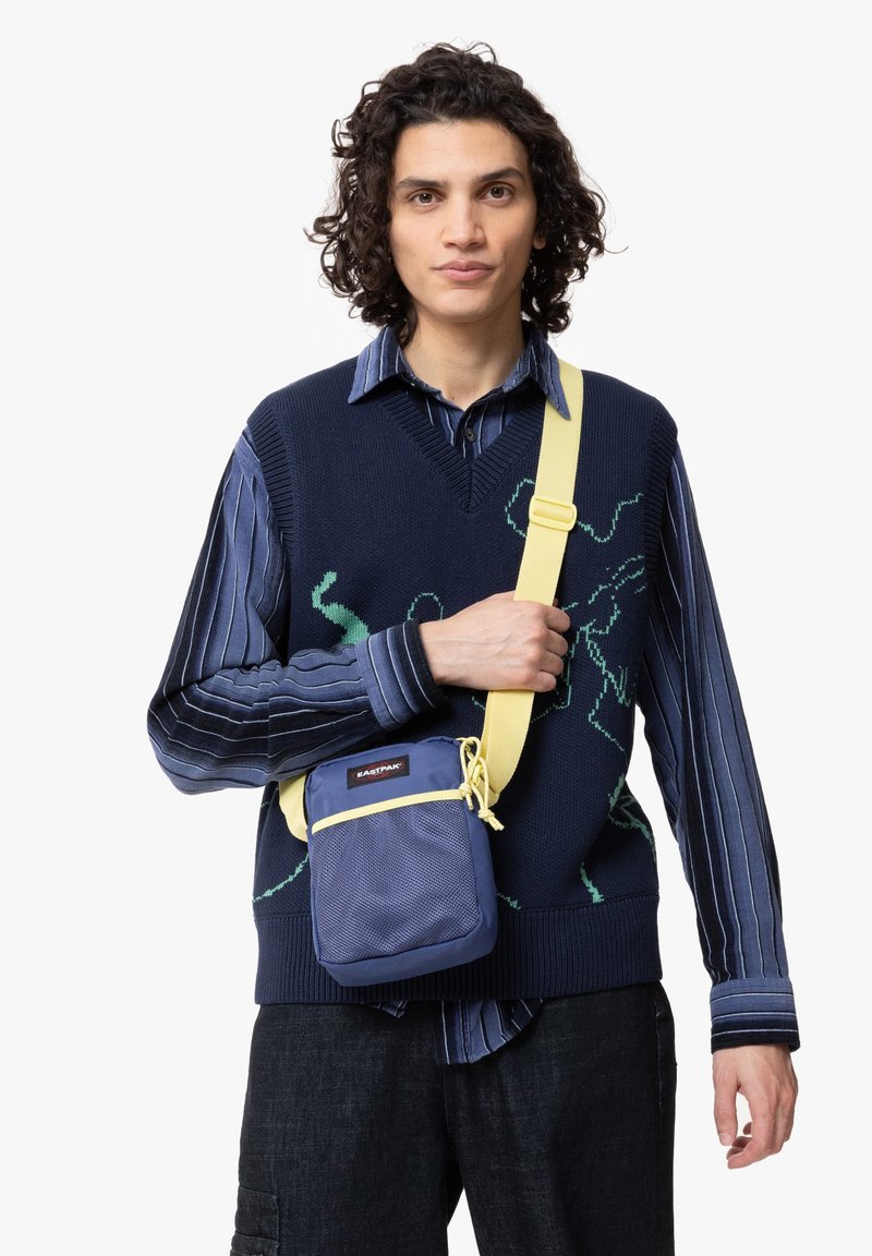 Mørkeblå crossbody taske med struktureret overflade, gul rem, justerbar spænde og frontlomme, der har et rødt mærkelogo.