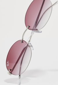 Lunettes de soleil Ray-Ban rondes avec des montures en argent fines et des verres teintés rose, présentées de biais avec des plaquettes de nez visibles sur un fond blanc.