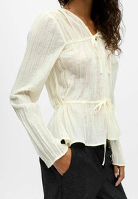 Blouse crème clair en tissu texturé, avec des rayures verticales, des manches longues, une taille froncée et une fermeture à nouer au cou.