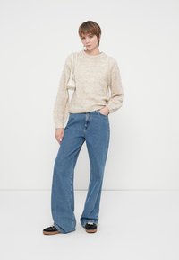 Krémszínű kötött pulóver textúrált mintával, magasan derékra szabt kék, széles szárú farmerral és fekete cipővel párosítva. Kis fehér táska.