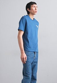 Mėlyna medvilnė t-shirt su trumpomis rankovėmis ir apvaliu kaklu, su mažu baltiu logotipu kairėje krūtinės pusėje, derinama su išblukusiomis mėlynomis džinsomis.