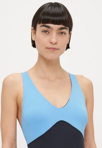 Μπλε και μαύρο μαγιό με σχέδιο σε σχήμα V, που διαθέτει μπλε μονόχρωμο πάνω μέρος και μαύρο κάτω πάνελ, κατασκευασμένο από λείο ελαστικό υλικό.