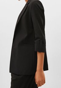 Blazer negro hecho de una tela suave, con un diseño ajustado, solapas muescadas y mangas enrolladas. Cuenta con bolsillos laterales y una forma entallada.