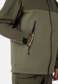 Olivgrüne Softshell-Jacke mit durchgehendem Reißverschluss und Taschen, mit strukturiertem Design und Klettverschluss-Akzenten an den Ärmelbündchen. RECCO-Logo auf dem Ärmel.