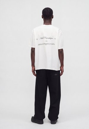 Personne debout, de dos, portant un t-shirt blanc avec texte imprimé et un pantalon noir ample sur un fond clair uni.