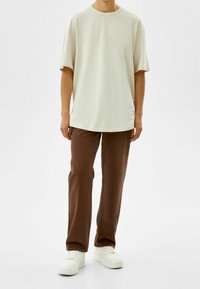Persona vistiendo una camiseta beige holgada, pantalones rectos marrones y zapatillas blancas, frente a un fondo claro y liso.