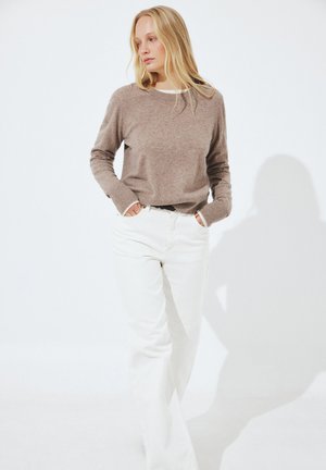 Donna con capelli biondi lunghi, che indossa un maglione color taupe e pantaloni bianchi, in piedi con le mani nelle tasche su uno sfondo bianco.