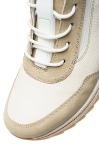 Sneaker with a beige suede upper and white leather accents. Features flat white laces and a textured toe box, with a rubber sole showing tread patterns.