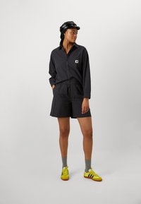 Svart skjorta med knappar och shorts i texturerad tyg, parat med gula Adidas-skor och grå strumpor. En platt svart keps framhäver stilen.