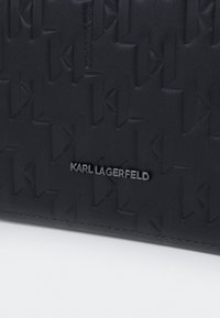 Superficie in pelle nera testurizzata con motivo geometrico in rilievo e piccolo logo metallico "Karl Lagerfeld" centrato vicino al bordo inferiore.