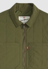 Veste matelassée olive avec fermeture éclair à l'avant, deux poches frontales, coutures contrastées et étiquette de marque à l'intérieur du col.