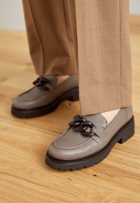 Gray leather loafers feature a chunky black chain accent, square toe, and thick rubber sole, paired with light brown trousers.