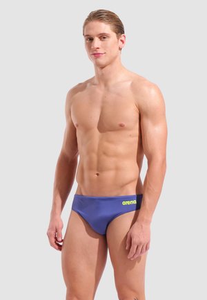 Joven con el pelo peinado hacia atrás que lleva unos calzoncillos de baño azules de Arena, de pie frente a un fondo liso de color gris claro.