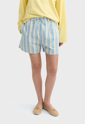 Osoba oblečená v svetlomodrých a béžových pruhovaných šortkách, žltom tričku s dlhým rukávom a béžových balerínach, stojaca s rukami v vreckách.