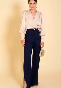 Top de satén rosa con mangas voluminosas, combinado con pantalones de tiro alto en navy con un botón dorado y aberturas laterales, complementado con un bolso de mano dorado.