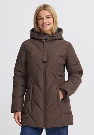 Femme souriante portant un manteau d’hiver matelassé marron avec capuche et fermeture éclair, mains dans les poches, debout devant un fond clair uni.