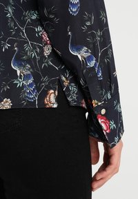 Camicia nera floreale con pavoni e fiori, in tessuto morbido, maniche lunghe e dettagli con bottoni sui polsini.