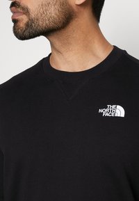 Homme barbu portant un sweat-shirt noir à col rond avec le logo blanc The North Face sur la poitrine gauche, regardant vers la gauche sur un fond uni.