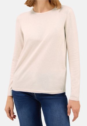 Femme portant un pull à manches longues beige clair et un jean bleu foncé, debout, les bras détendus le long du corps.