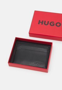 Black leather card holder displayed in a red box. Features multiple card slots and embossed logo. Smooth texture and minimal design.