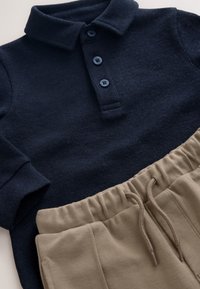 Pull-over bleu marine en tissu texturé avec patte de boutonnage à trois boutons, associé à un short beige à taille élastique, poches latérales et cordon de serrage.