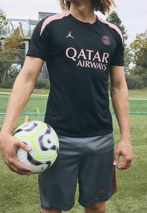 Persoon in een zwart-roze Paris Saint-Germain shirt houdt een witte en gele voetbal vast op een groen buitenveld.