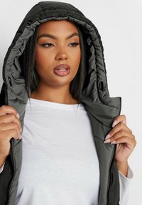 Svart vadderad väst med huva, med ett quiltat mönster, dragkedjestängning och snördetalj. Modellen håller den öppna fronten.