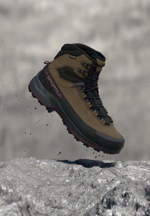 Botte de randonnée marron et noire avec des lacets rouges en l’air au-dessus d’une surface rocheuse, montrant une semelle robuste et des détails de design extérieur.