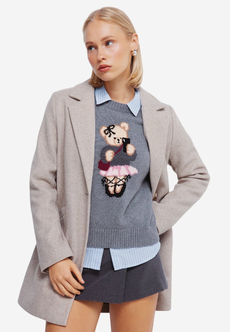 Grijze gebreide trui met een berenontwerp en een zwarte strik, gelaagd over een lichtblauw gestreept shirt en gecombineerd met een beige jas.