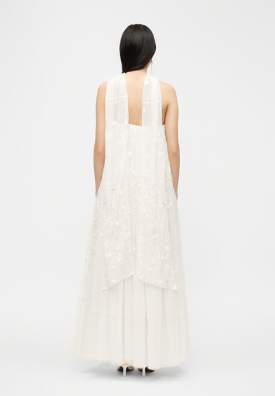 Pikkade tumedate juustega naine kannab valget varrukateta kleiti, millel on lillelise pitsiga pealiskate ja ristkülikukujuline madal seljatükk, ning seisab valgetel kontsadel tavalise tausta ees.