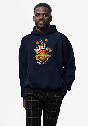 Mand iført navyblå hættetrøje med farverige Space Jam-tegneseriefigurer og basketballgrafik, kombineret med sorte bukser med hvide gitterlinjer.