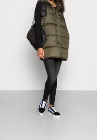 Osoba oblečená v olivově zelené péřové vestě, černém dlouhém rukávu, černých skinny džínech, iridescentních černých teniskách a nesoucí černou tašku.