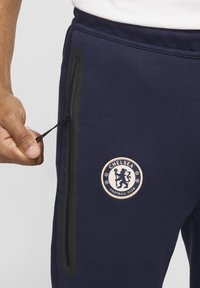 Laivonsiniset jumppahousut mustalla vetoketjullisella taskulla. Edessä on pyöreä beigenvärinen Chelsea Football Clubin logo. Pehmeä materiaali, joustava vyötärö.