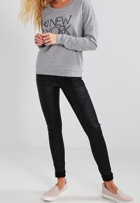 Persona che indossa un felpa grigia con la scritta "New York", leggings neri testurizzati e scarpe slipper rosa chiaro, in piedi contro uno sfondo semplice.