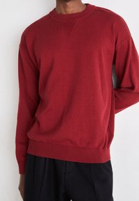 Roter Strickpullover mit rundem Halsausschnitt, gerippten Bündchen und Saum. Verfügt über ein strukturiertes Paneel auf der Brust für zusätzlichen Detail.