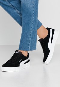 Černé semišové tenisky s bílými akcenty, plochými tkaničkami a silnou gumovou podrážkou. Úzký design s logem na jazyku a na boku.